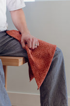 Person sitting on a wooden bench with a red patterned cloth draped over their lap against a plain background
